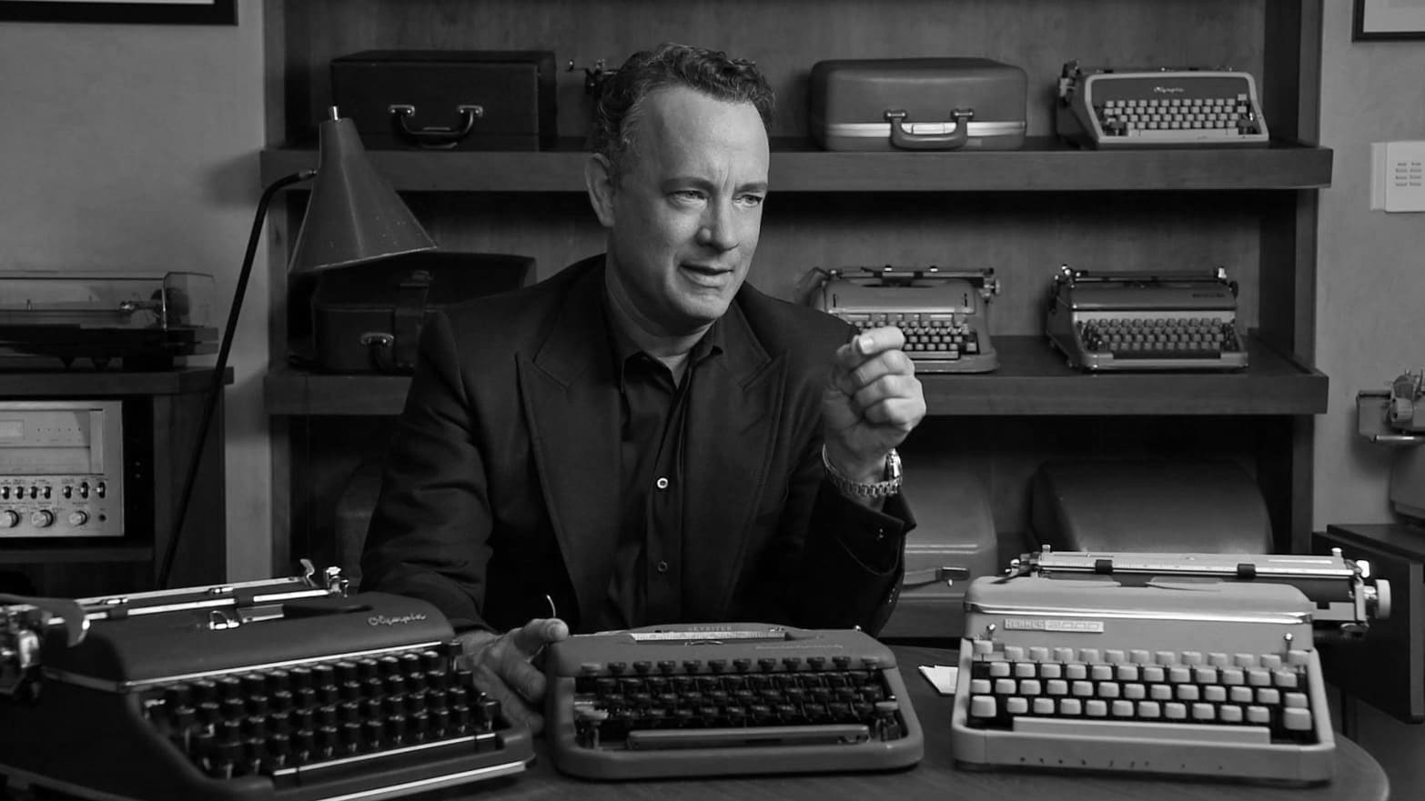 A man sits at a wooden desk surrounded by various vintage typewriters. He is captured in mid-conversation with expressive hand gestures. Behind him, more typewriters are neatly arranged on shelves. The image is in black and white, giving it a classic, nostalgic feel.