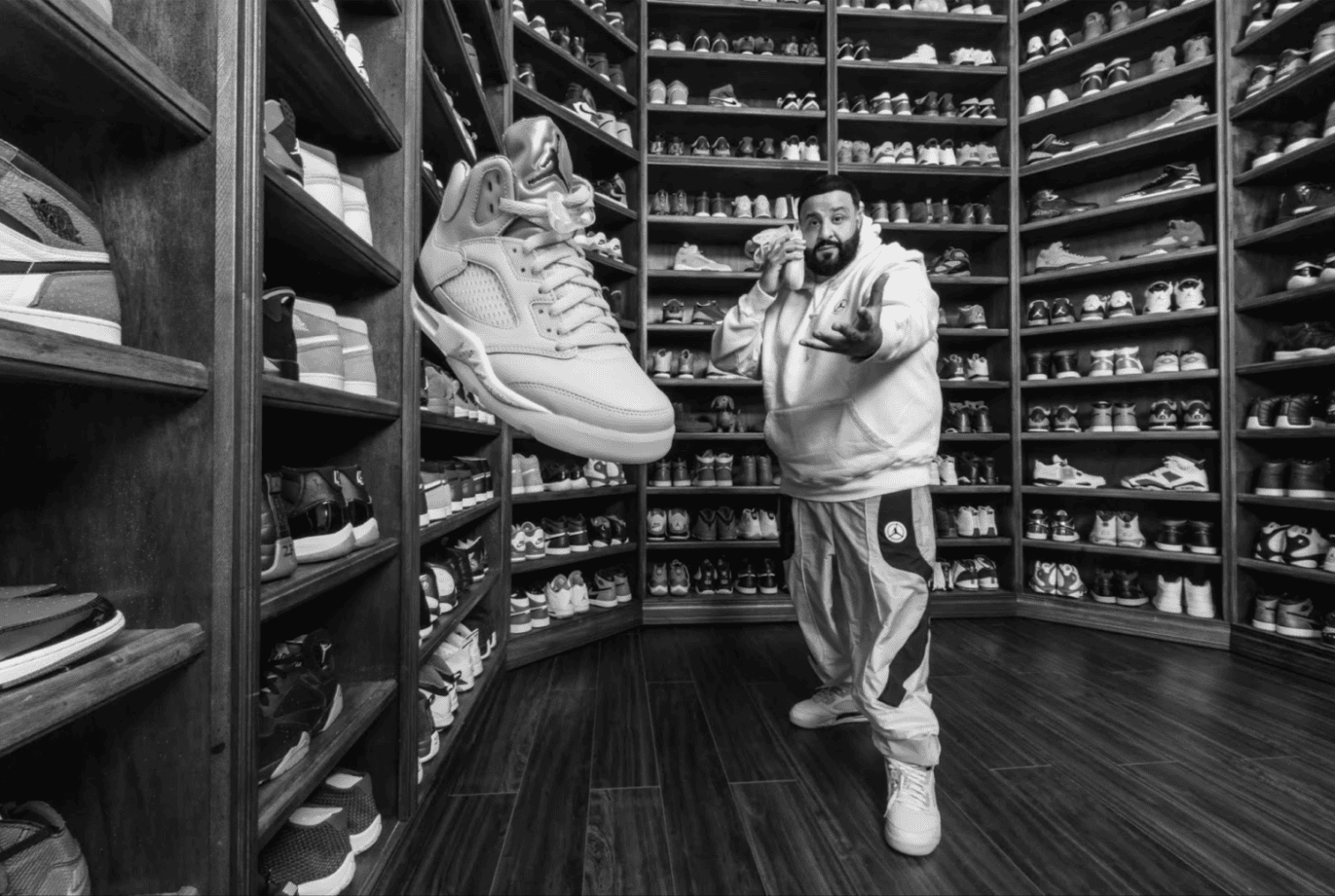 A man stands in a room surrounded by shelves filled with sneakers. He points towards a large floating sneaker in the foreground. The scene is monochromatic, highlighting the extensive sneaker collection and the man's dynamic pose.