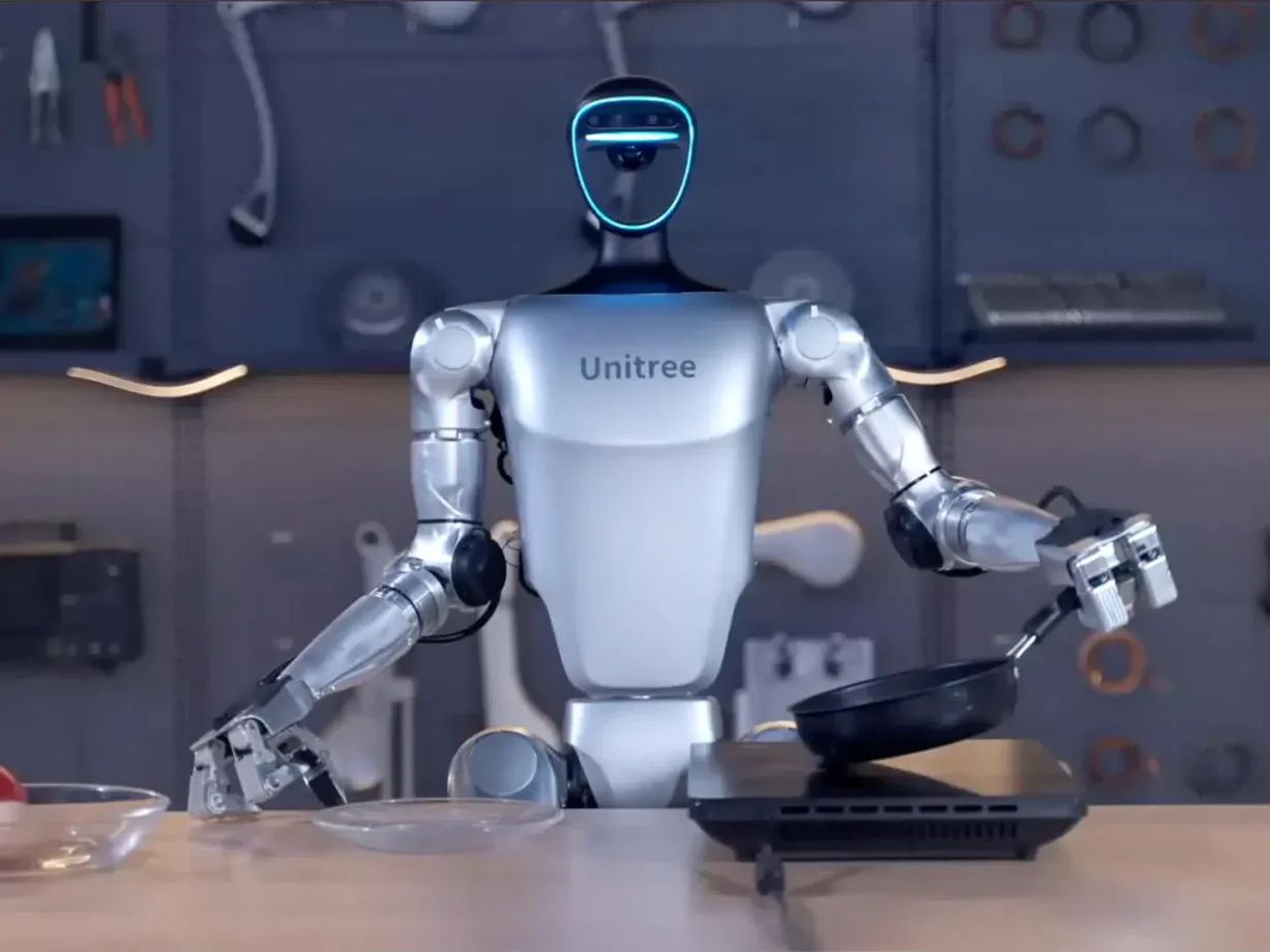 A humanoid robot with "Unitree" on its chest is standing in a kitchen-like setting, holding a frying pan in one hand and a spatula in the other. It appears to be cooking, with various tools visible in the background.