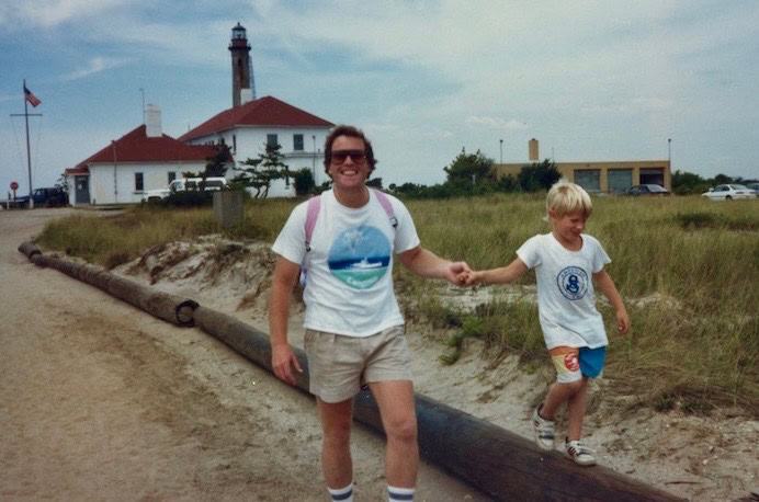 An adult and a child hold hands and walk along a log path near grassy dunes. A lighthouse and building with an American flag are visible in the background under a partly cloudy sky.