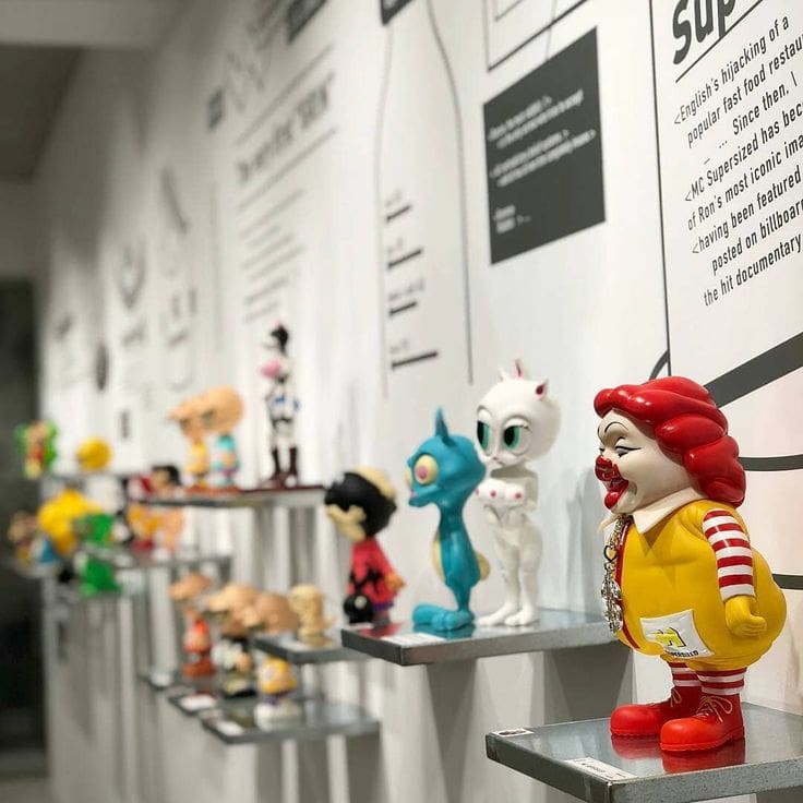 Colorful figurines, including a clown in a yellow suit, are displayed on individual shelves against a white wall with text and graphics, creating a playful and modern exhibition.