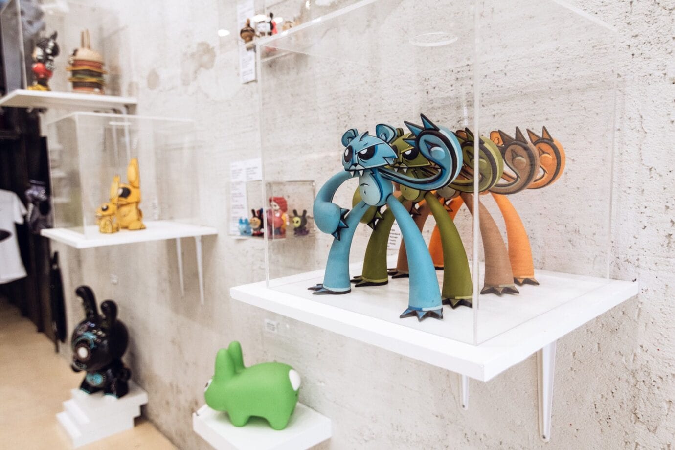 A display of colorful, stylized animal figurines and designer toys are enclosed in transparent boxes on white shelves mounted to a concrete wall in a gallery or shop setting.