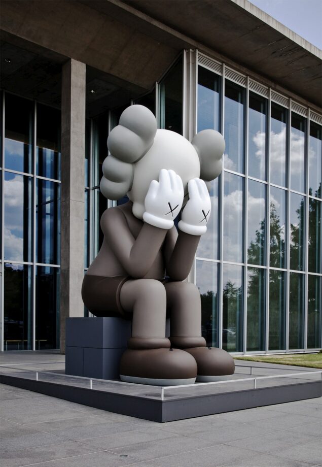 A large sculpture of a cartoon-like figure with gray skin, white gloves, and "X" eyes sits on a bench with its face buried in its hands, positioned outside a modern glass building.