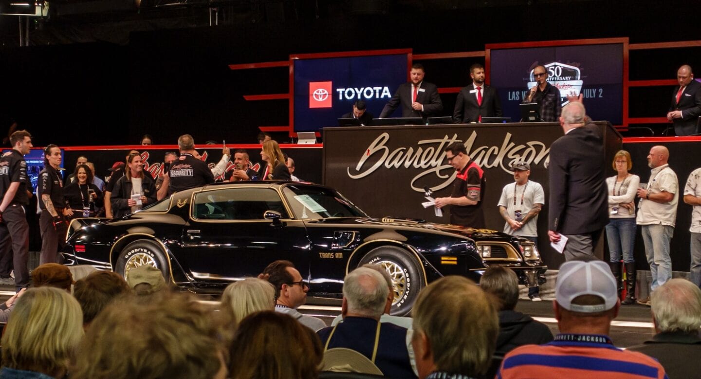A classic black Pontiac Trans Am is showcased on an auction stage, surrounded by a crowd. Auctioneers and attendees look on, with the Barrett-Jackson logo and a Toyota sign visible in the background.