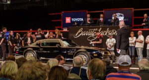A classic black Pontiac Trans Am is showcased on an auction stage, surrounded by a crowd. Auctioneers and attendees look on, with the Barrett-Jackson logo and a Toyota sign visible in the background.