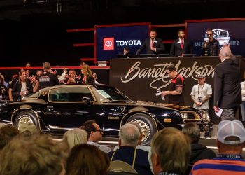 A classic black Pontiac Trans Am is showcased on an auction stage, surrounded by a crowd. Auctioneers and attendees look on, with the Barrett-Jackson logo and a Toyota sign visible in the background.